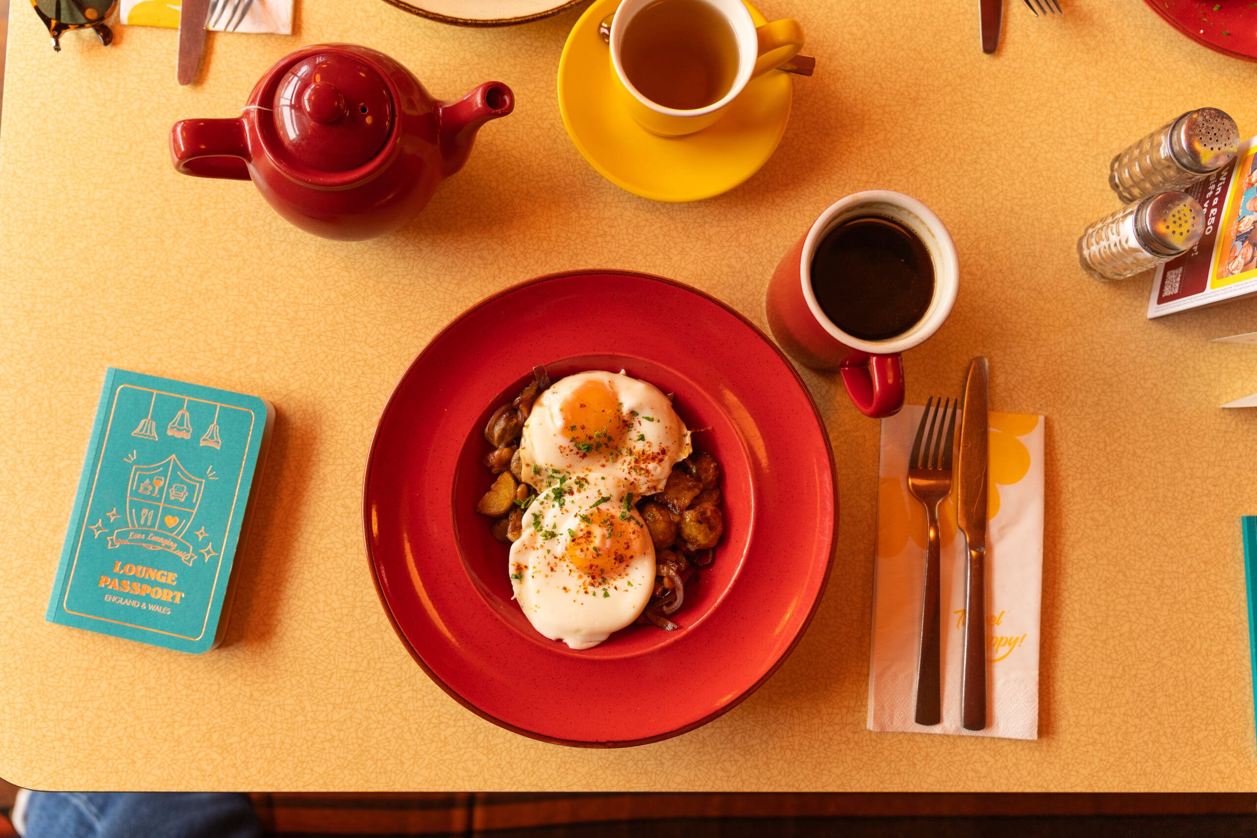 A plate with eggs over roasted potatoes and vegetables sits on a table with a red teapot, two coffee cups, utensils, a blue passport-style booklet, and salt and pepper shakers.