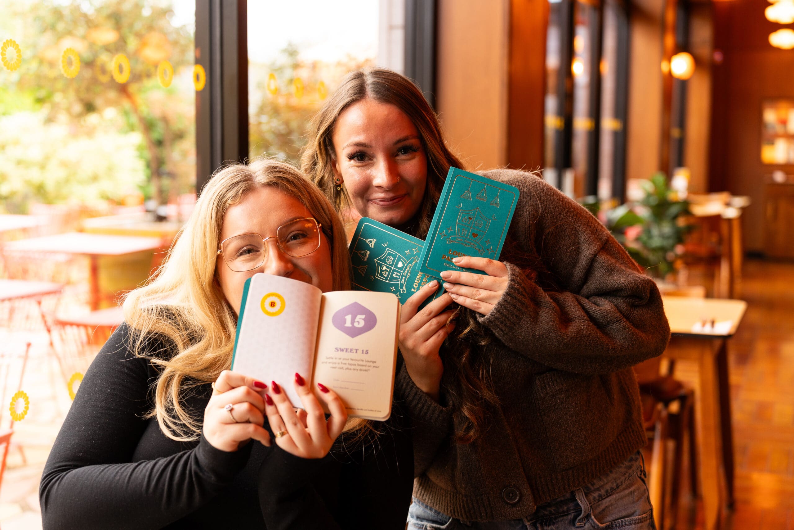 Two women sit in a cozy café by a window, smiling and holding open colorful books towards the camera. Warm lighting and outdoor scenery create a cheerful, inviting atmosphere.