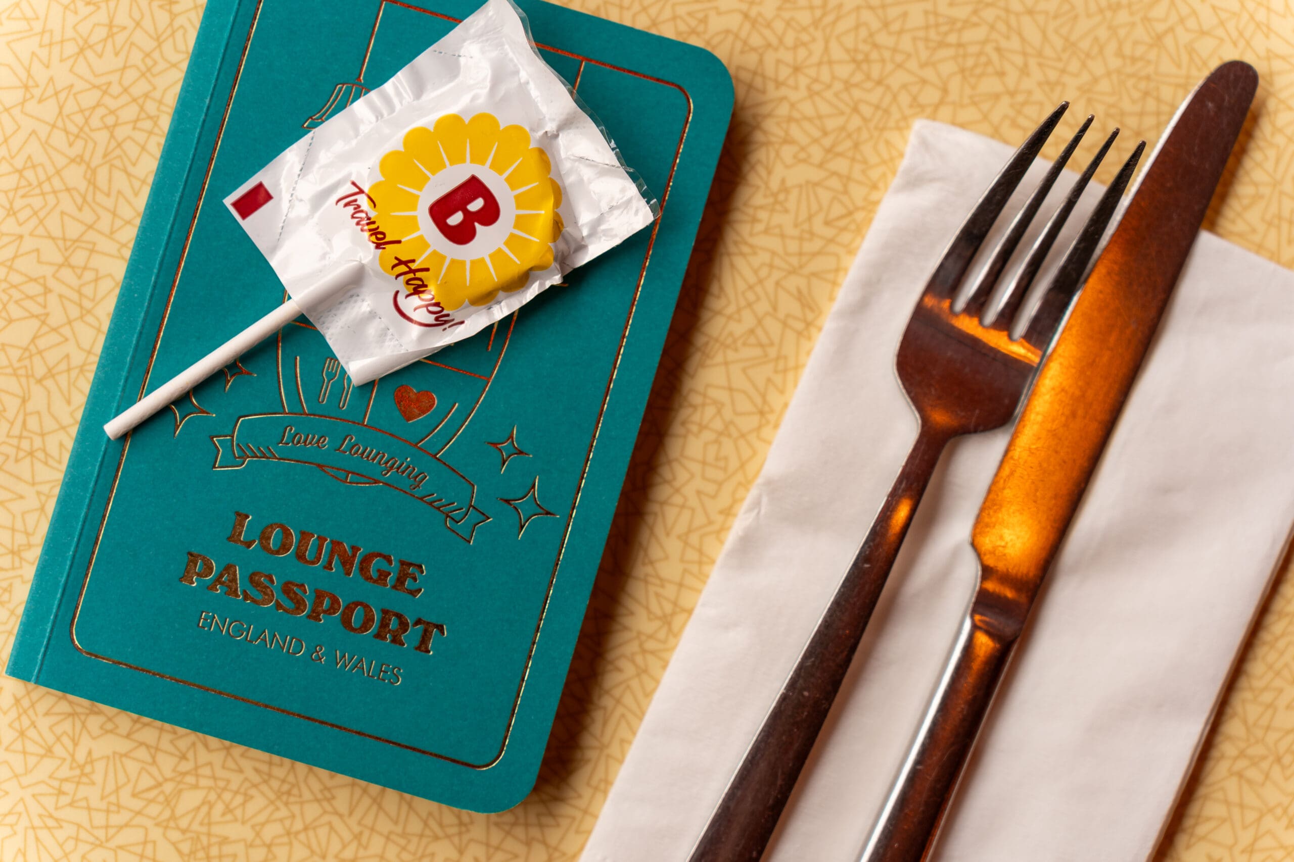A teal "Lounge Passport" booklet with a lollipop on top sits next to a fork and knife placed on a white napkin, all on a yellow textured surface.