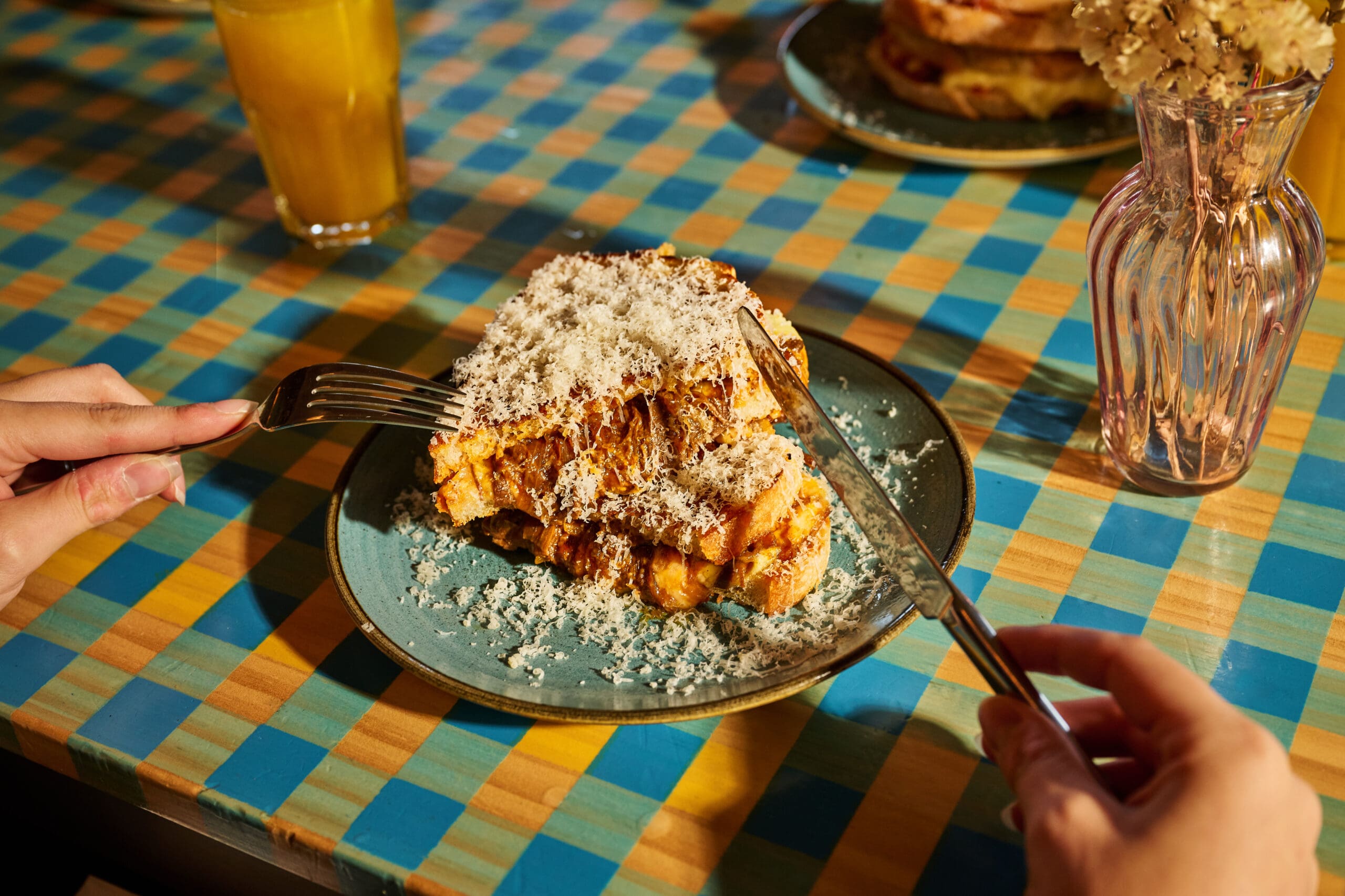 A person uses a knife and fork to cut into thick slices of bread topped with grated cheese on a blue plate, set on a colorful checkered tablecloth. A glass of orange juice and a vase of dried flowers are nearby.