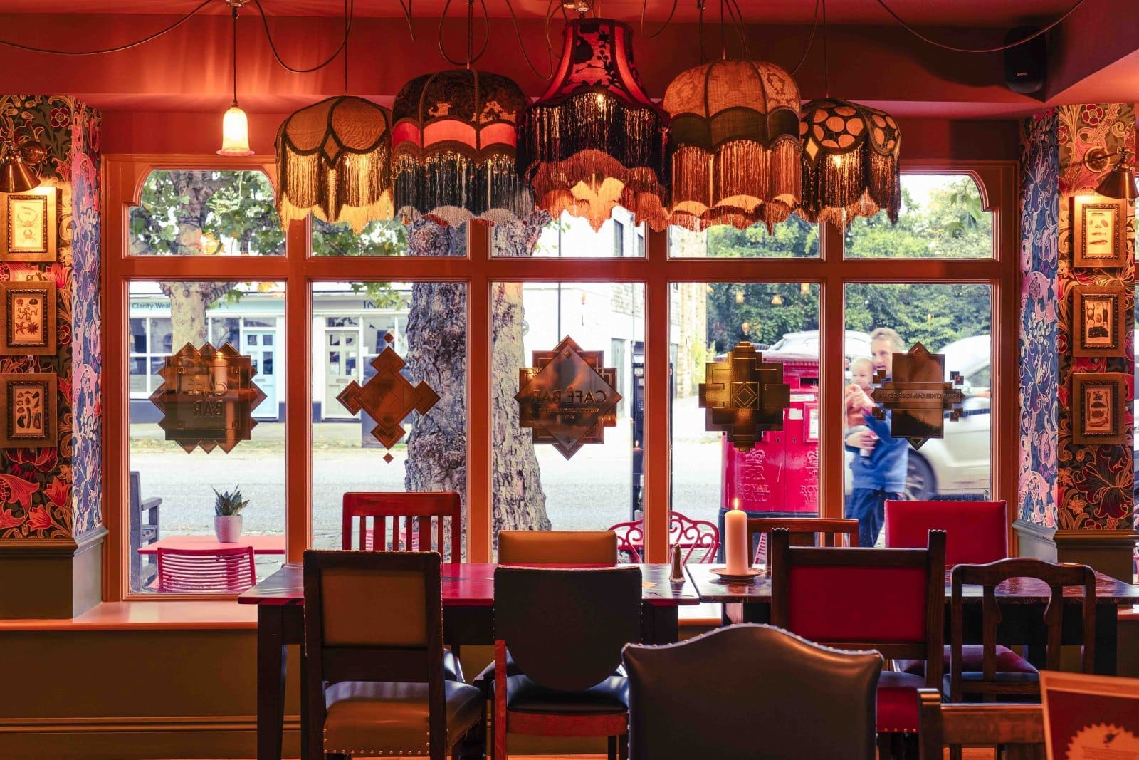 A cozy Bruto café interior with vintage lampshades hanging above mismatched chairs and tables. Large windows reveal trees, a red post box, and a person walking outside. Patterned wallpaper and framed art decorate the walls.