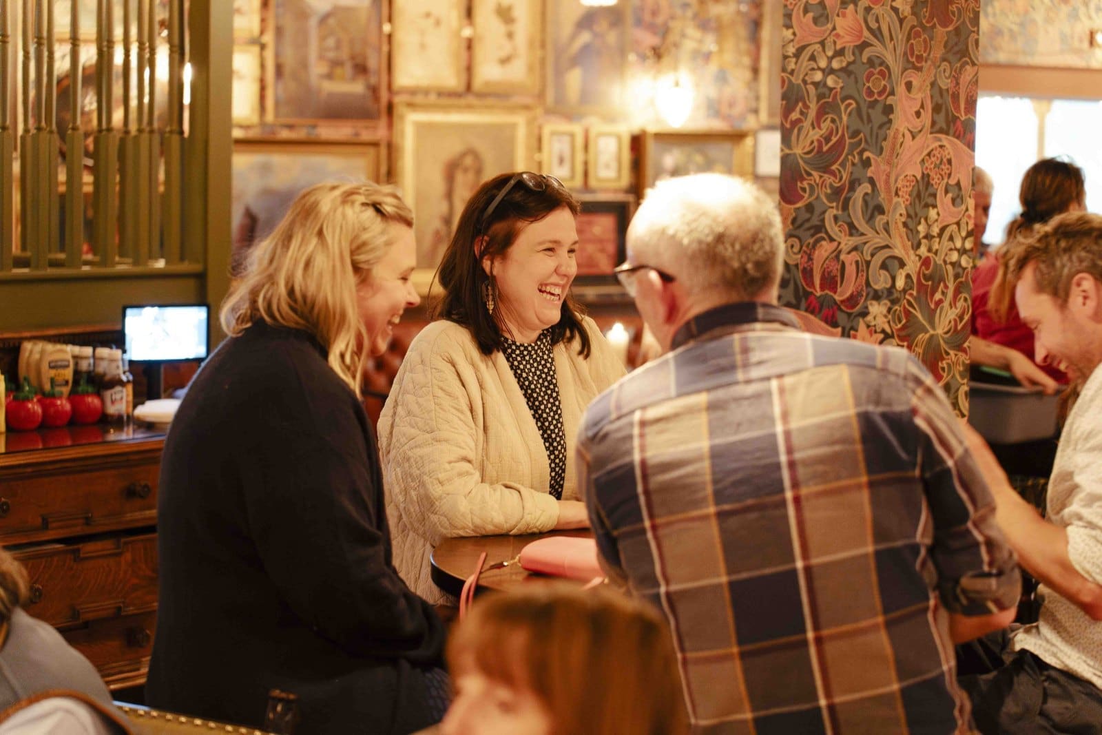 Four people sit around a table in a cozy, warmly lit Bruto café, smiling and talking. The room features patterned wallpaper, framed pictures on the walls, and a rustic, inviting atmosphere.
