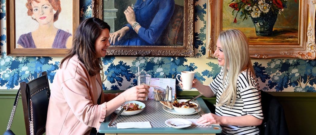 Two women sit at a small table in Caballero cafe, smiling and holding drinks. Colorful portraits and a flower painting hang on a blue-patterned wall behind them. Food and tableware are arranged on the table.