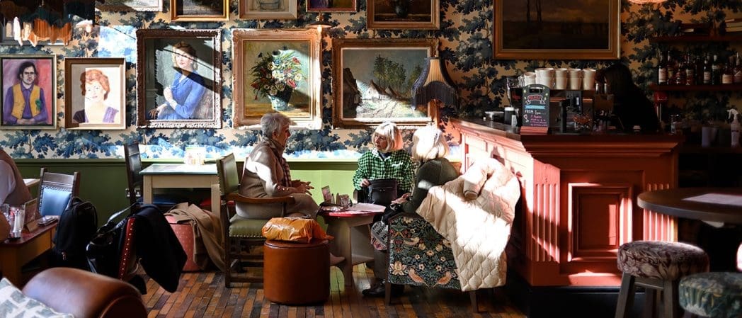 Two elderly women sit and chat on a couch in a cozy, Caballero-inspired café, with sunlight streaming in, surrounded by framed paintings, vintage furniture, and a warmly lit bar area.