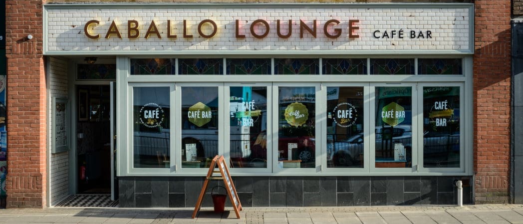Storefront of "Caballo Lounge Café Bar" with large windows, Caballo-themed decorative stained glass, and a sidewalk sandwich board sign in front. The reflection of parked cars is visible in the window.