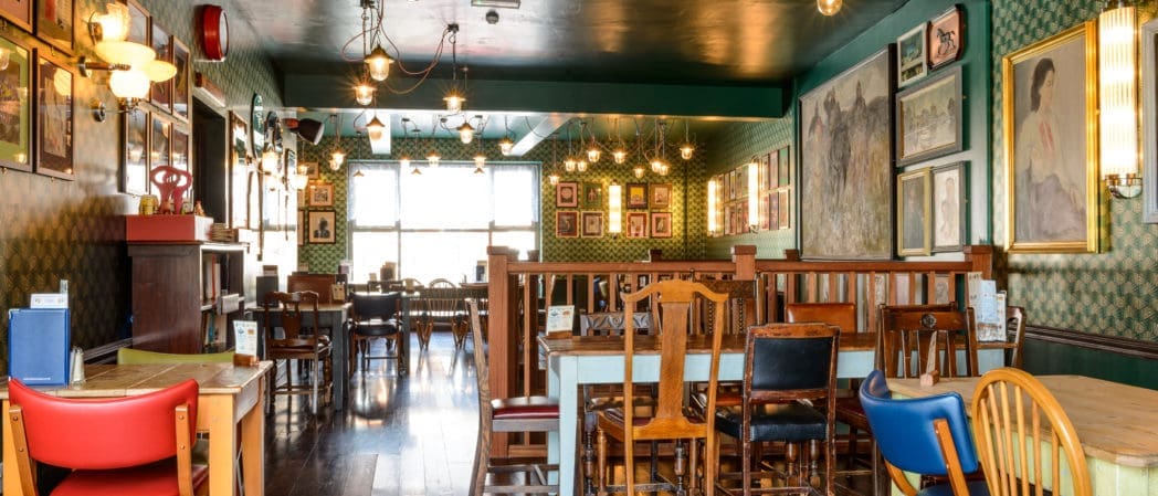 A cozy restaurant with vintage decor, mismatched wooden chairs and tables, green patterned wallpaper, framed artwork—including a striking caballo painting—and warm lighting. Large windows let in natural light.