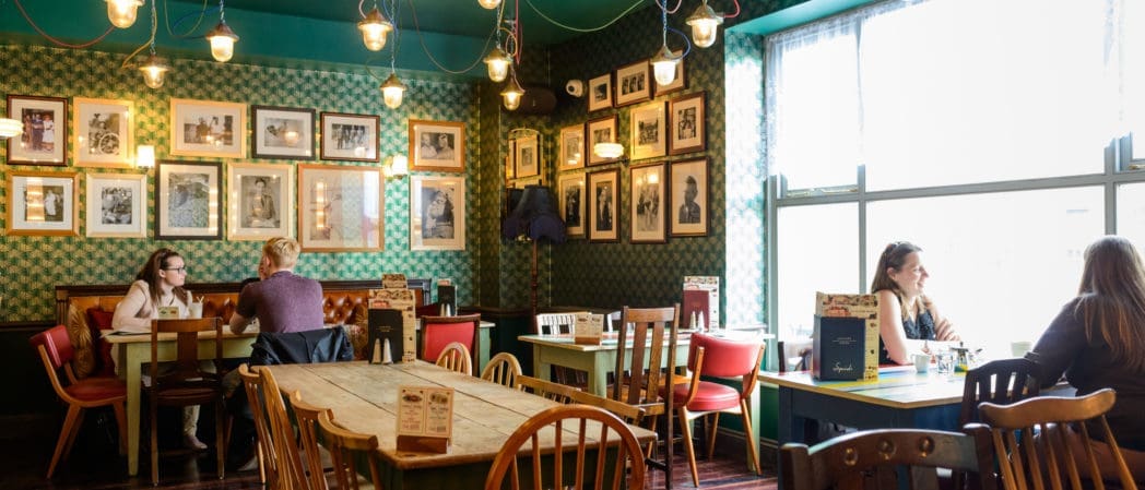 Cozy café with patterned green walls, framed photos—including one of a caballo—mismatched wooden chairs, and hanging lights. A few people sit at tables chatting near large windows with natural light streaming in.