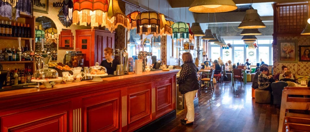 A cozy café with colorful lampshades, a red wooden counter, cakes and drinks on display, and people chatting at tables. Warm lighting and eclectic decor—complete with a playful Caballo figurine—create a welcoming atmosphere.