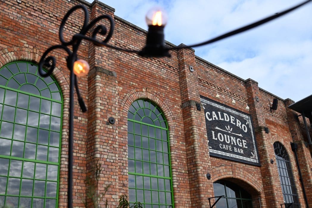 A brick building with large arched windows and a sign that reads "Caldero Lounge Café Bar." String lights hang in the foreground, creating a warm, inviting glow at Caldero against a partly cloudy sky.