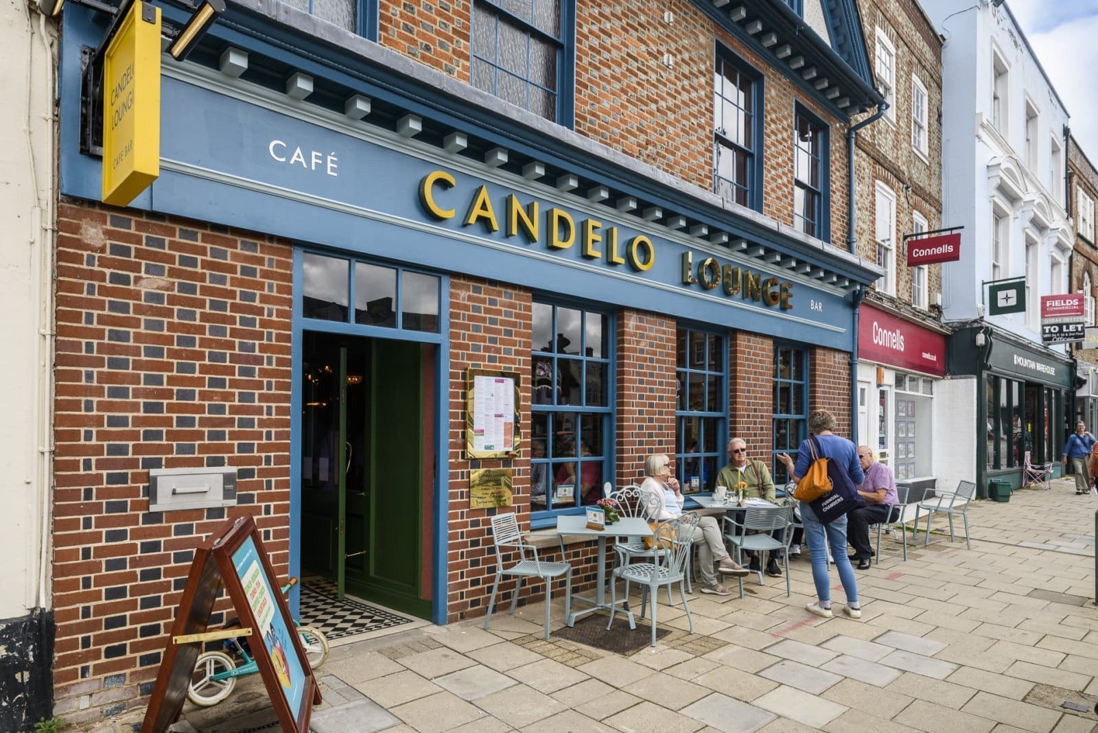 People sit at tables outside Candelo Lounge café on a brick-paved street lined with shops. The café’s blue and yellow Candelo sign, large windows, and lively customers create a welcoming spot for drinks and conversation.