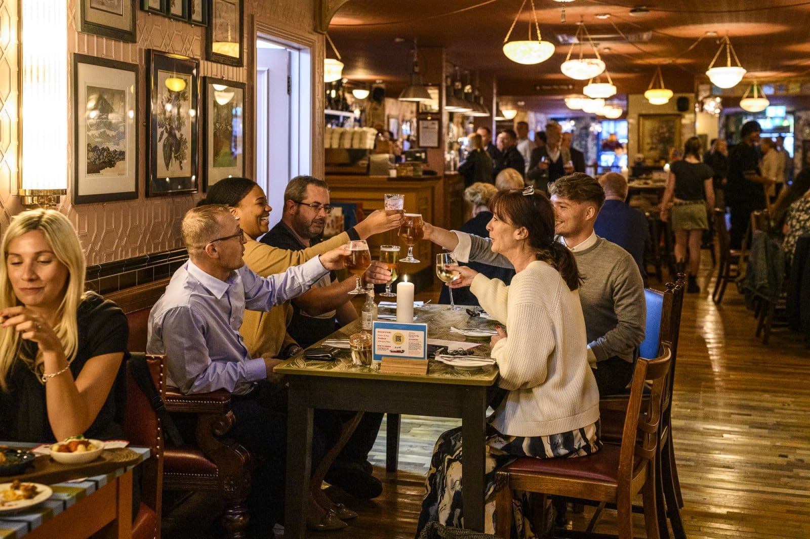 A group of people sit around a restaurant table in Candelo, raising their glasses for a toast. Other diners and warm lighting create a busy, lively atmosphere in the background.