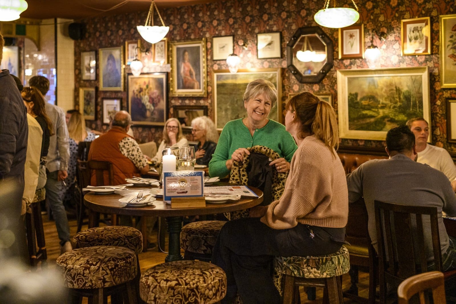 Two women sit and laugh at a round table in Candelo, a cozy, warmly lit restaurant decorated with patterned wallpaper and framed art. Other diners chat and eat in the background, creating a lively, friendly atmosphere.