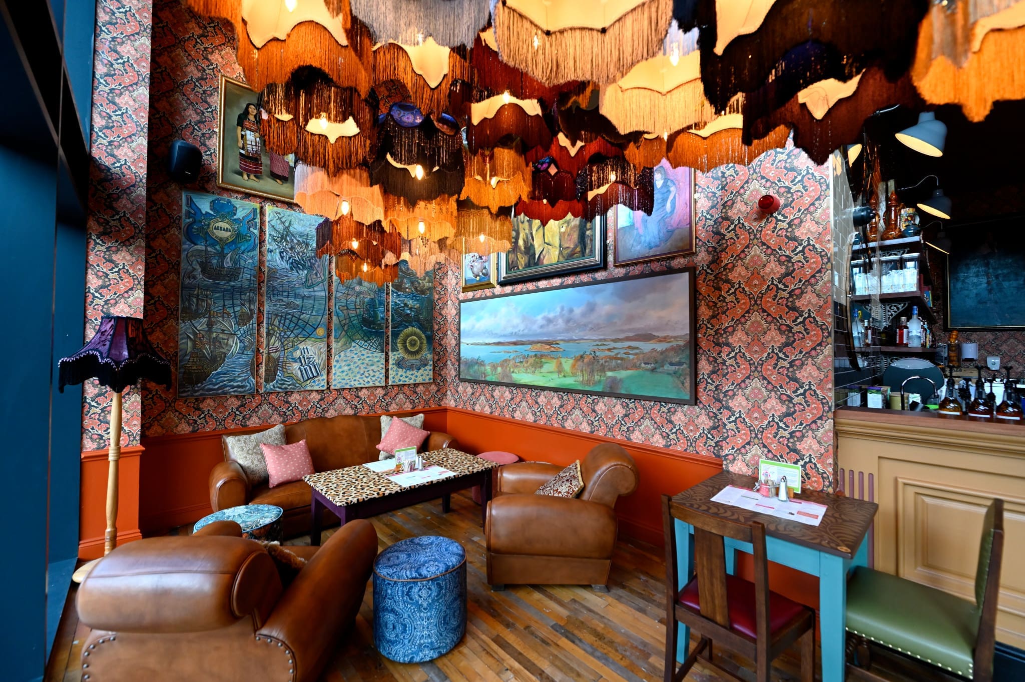 Eclectic cafe interior with leather chairs, patterned wallpaper, colorful lampshades hanging from the ceiling, various framed paintings by Carlo, and cozy seating around low tables on a wooden floor.