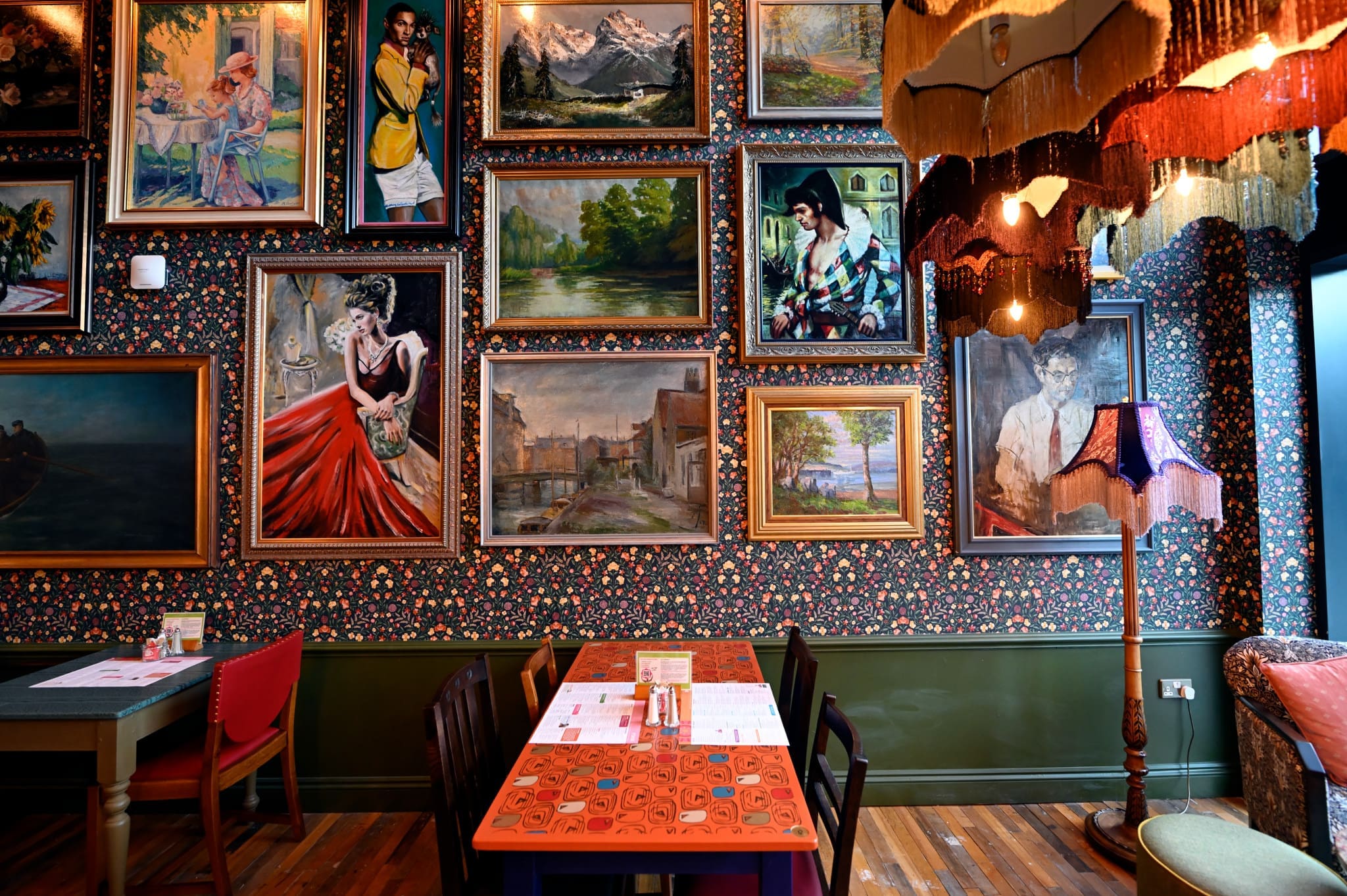 A cozy restaurant interior by Carlo, with patterned wallpaper, eclectic framed paintings covering the wall, colorful chairs around wooden tables with menus, and vintage-style lamps providing warm light.