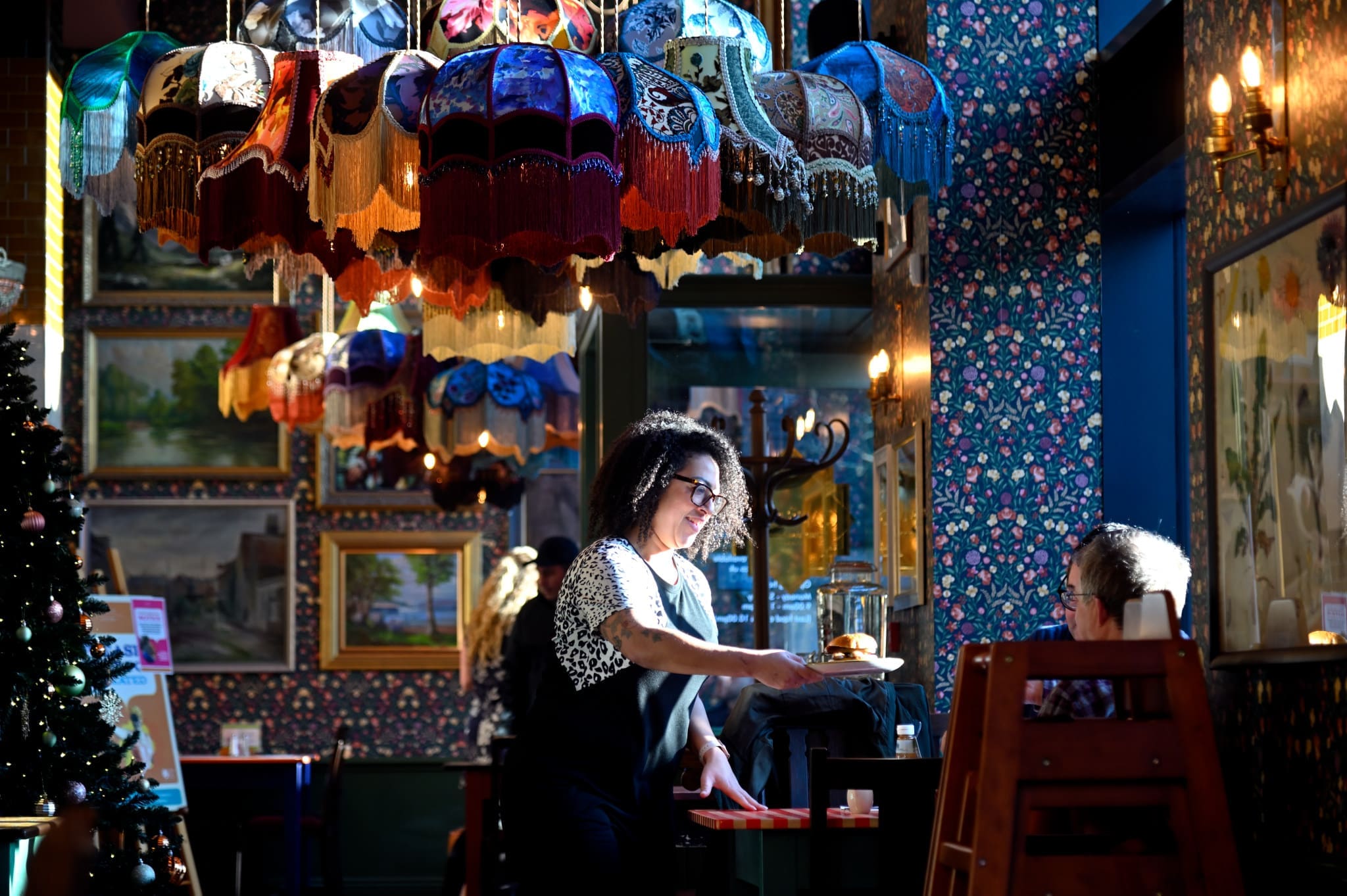 A smiling waitress serves food to a seated customer in Carlo’s cozy, colorful restaurant with vintage lamps, floral wallpaper, and framed paintings on the walls. Sunlight streams in from a nearby window.