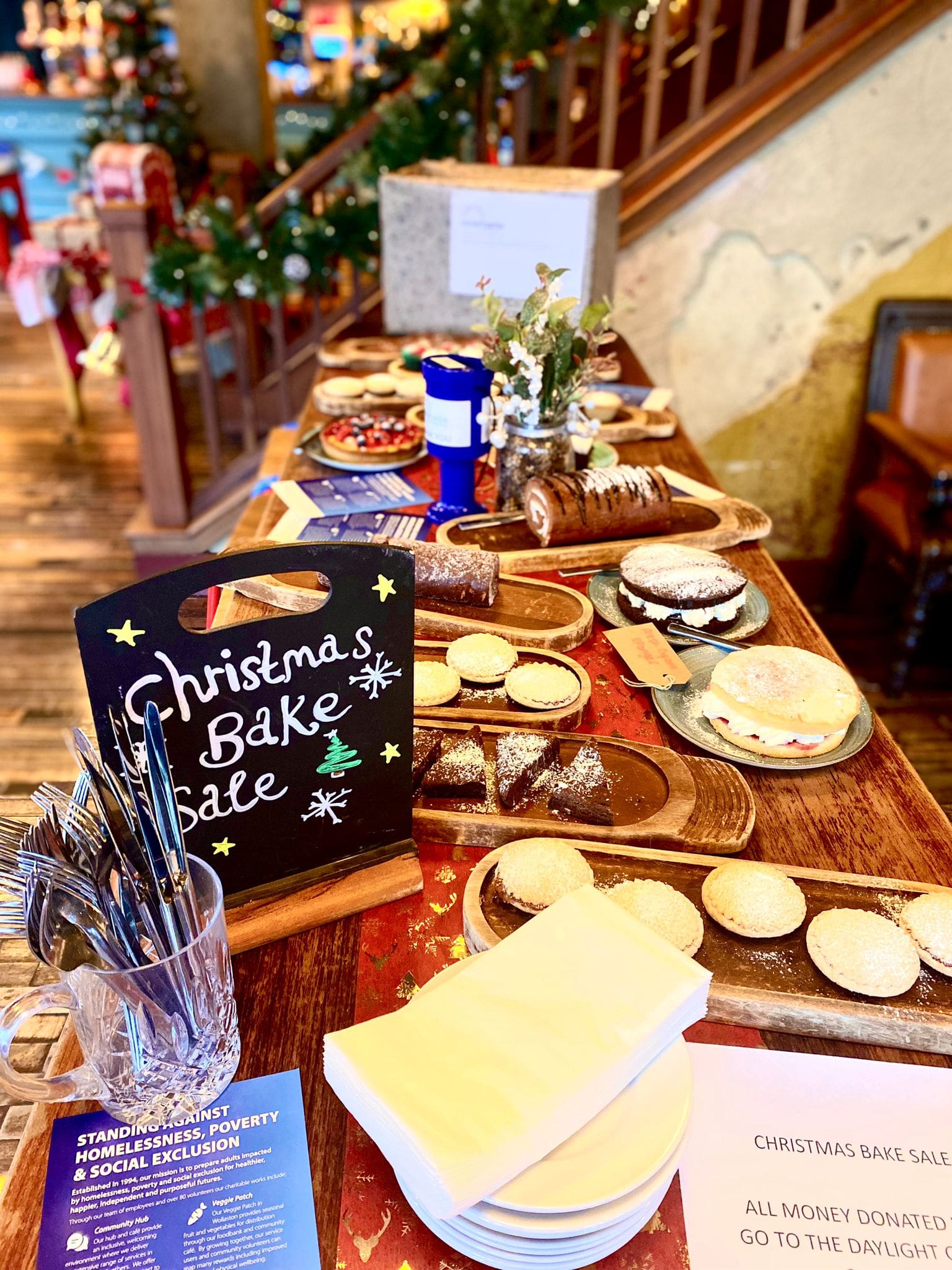 A festive table displays various baked goods for a Christmas bake sale, with mince pies, yule logs, and cakes. A chalkboard sign reads “Christmas Bake Sale” beside donation flyers and utensils. Holiday decorations are visible in the background.