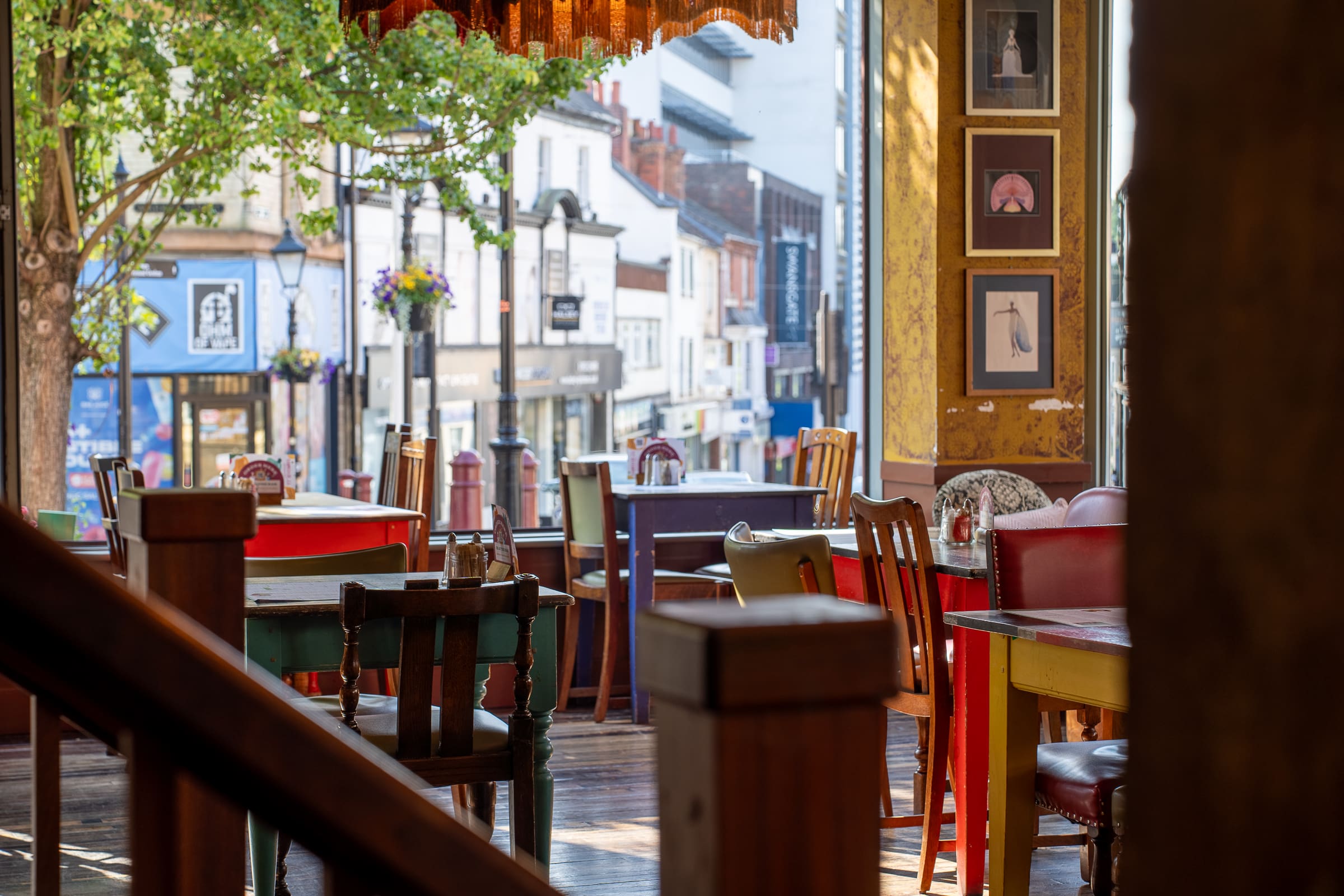 A cozy café with colorful mismatched chairs and tables sits empty near large windows, offering a view of Castello’s lively street with shops and trees. Framed pictures decorate the yellow walls, adding charm to this inviting spot.