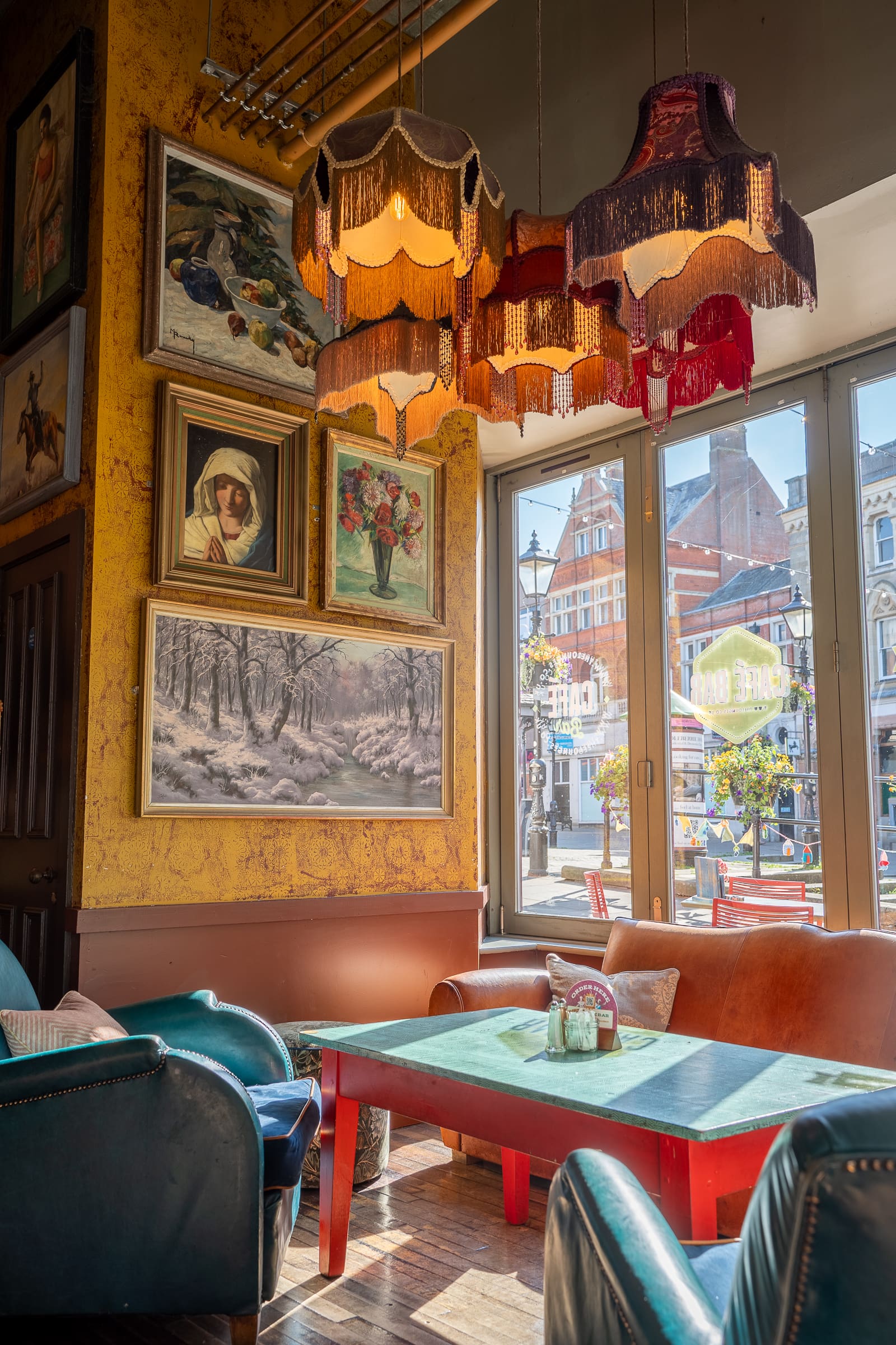 Cozy cafe interior with vintage armchairs, a green table, colorful fringed lamps, and walls decorated with various framed paintings. Large windows let in sunlight and offer a Castello view of the lively street outside.
