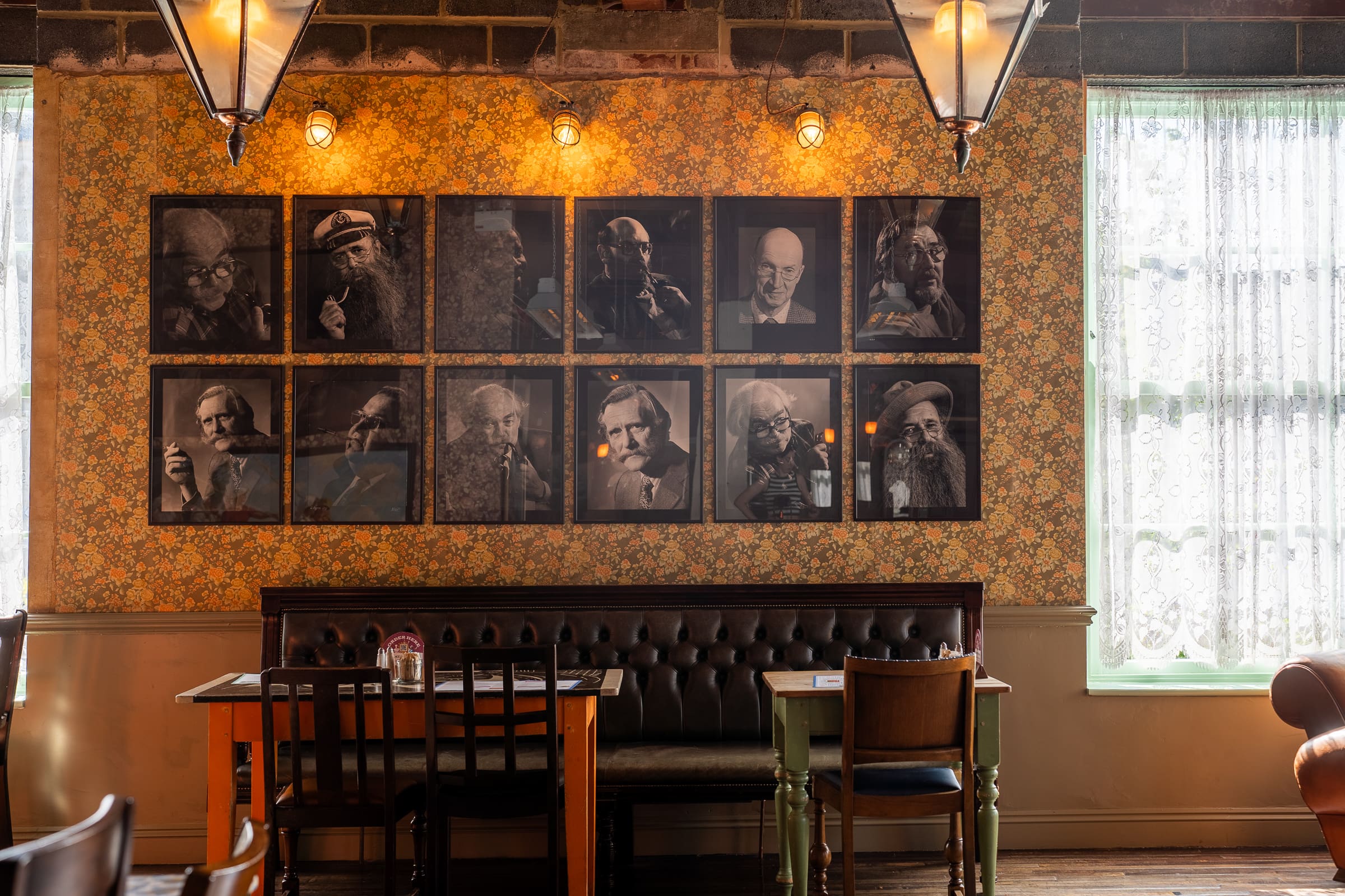 A cozy cafe with vintage wallpaper, framed portraits, and wooden tables offers a Castello charm. Relax on a bench under warm hanging lights near tall windows with lace curtains, soaking in an inviting and nostalgic atmosphere.