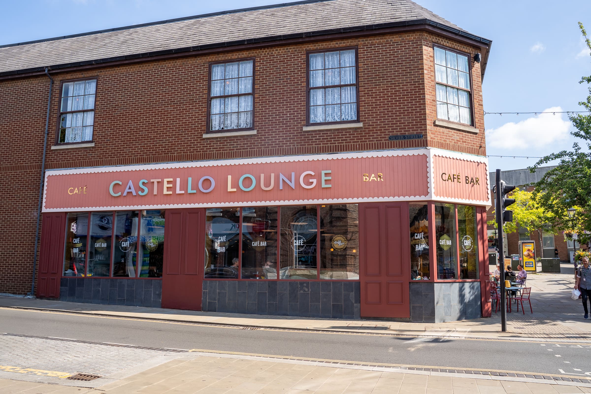 Castello Lounge, part of the Castello family, is a café bar with large windows and a red and cream storefront, nestled on the corner of a brick building along a quiet street with a few people relaxing outside.