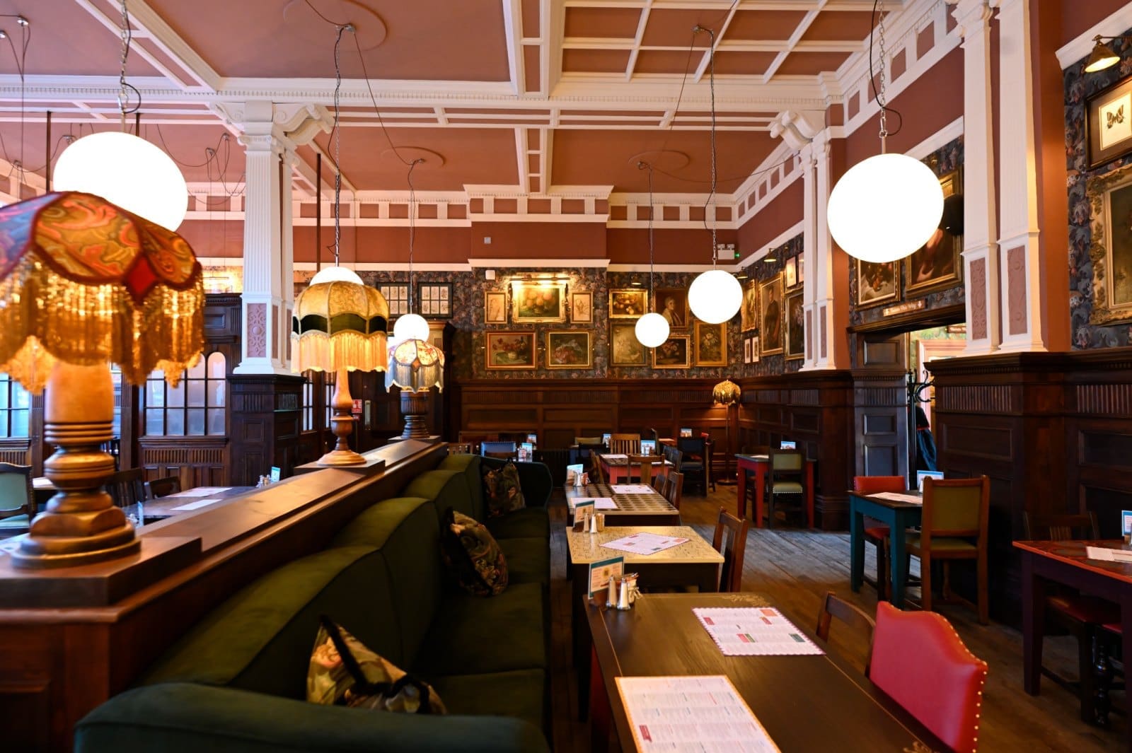 A cozy restaurant interior with vintage lamps, round hanging lights, dark wooden paneling, green upholstered seating, and framed artwork on the walls exudes centivo charm. Tables are set and the space appears inviting and warmly lit.