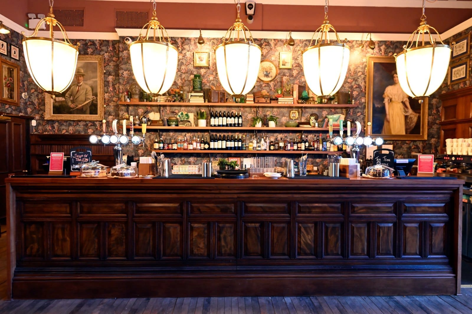 A stylish bar with a dark wood counter, large pendant lights above, shelves lined with bottles and glasses, floral wallpaper, and framed portraits on the walls. Centivo-branded pastries and menus are displayed on the counter.