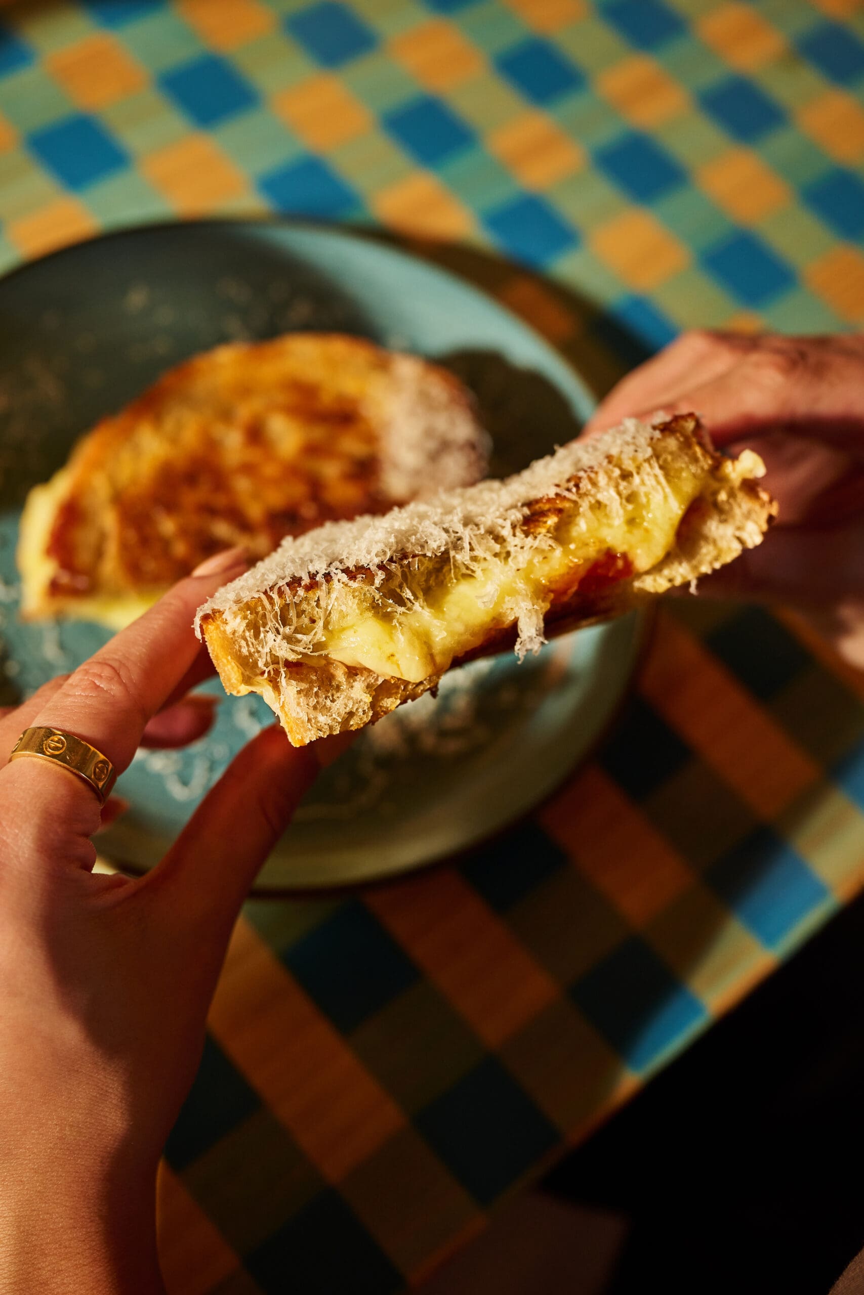 A close-up of hands holding a half-eaten grilled cheese sandwich with melted cheese inside, above a plate on a colorful checkered tablecloth.