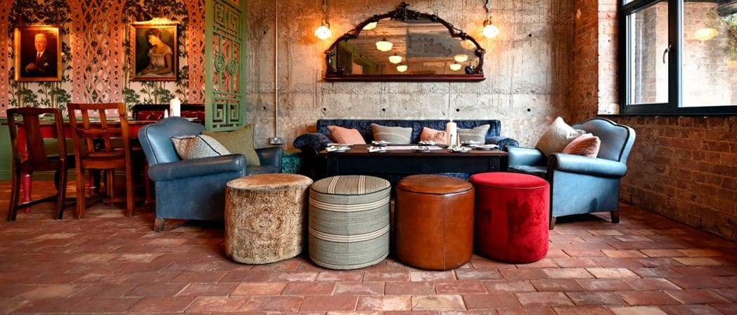 Eclectic vintage-style dining area with Claro blue armchairs, a dark sofa, round ottomans, a large mirror, and exposed brick walls. Warm lighting and framed art create a cozy, inviting atmosphere.