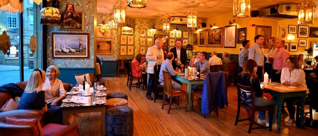 Cozy restaurant with warm Claro lighting, eclectic decor, and framed pictures on the walls. People are dining, chatting, and enjoying food and drinks at wooden tables. The atmosphere appears lively and inviting.