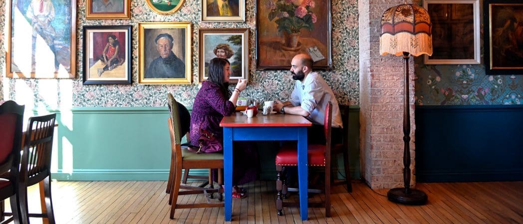 A man and woman sit across from each other at a colorful table in Claro, a cozy, vintage-style café decorated with floral wallpaper, framed portraits, and a tall, ornate floor lamp.