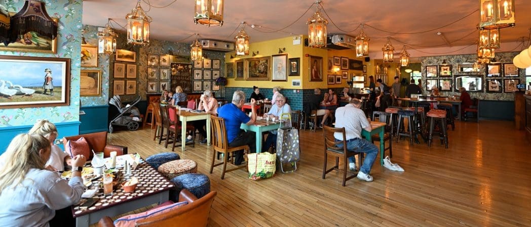 A cozy, well-lit restaurant with wooden floors, eclectic artwork on the walls, and several groups of people dining at various tables. Warm hanging lanterns create a relaxed atmosphere, making Claro an inviting place to gather.