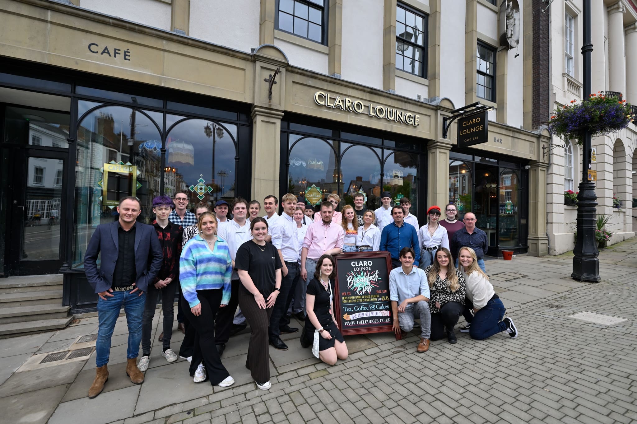 A group of people pose and smile outside Claro Lounge café. They stand and kneel on the stone pavement near a sign displaying the Claro name, with large windows and hanging flower baskets decorating the vibrant building.
