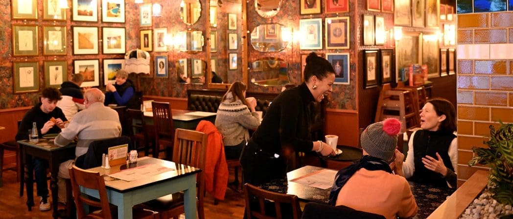 A cozy, warmly lit restaurant with framed artwork covering the walls. Diners sit at tables enjoying conversations, while a smiling Clavio waitress serves drinks to two women at a table in the foreground.