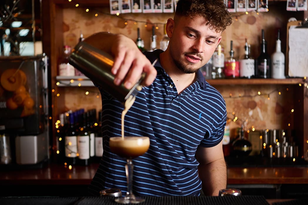 A bartender in a blue striped polo shirt, sporting a Bonzo grin, pours a creamy drink from a shaker into a cocktail glass at a bar decorated with fairy lights and bottles in the background.