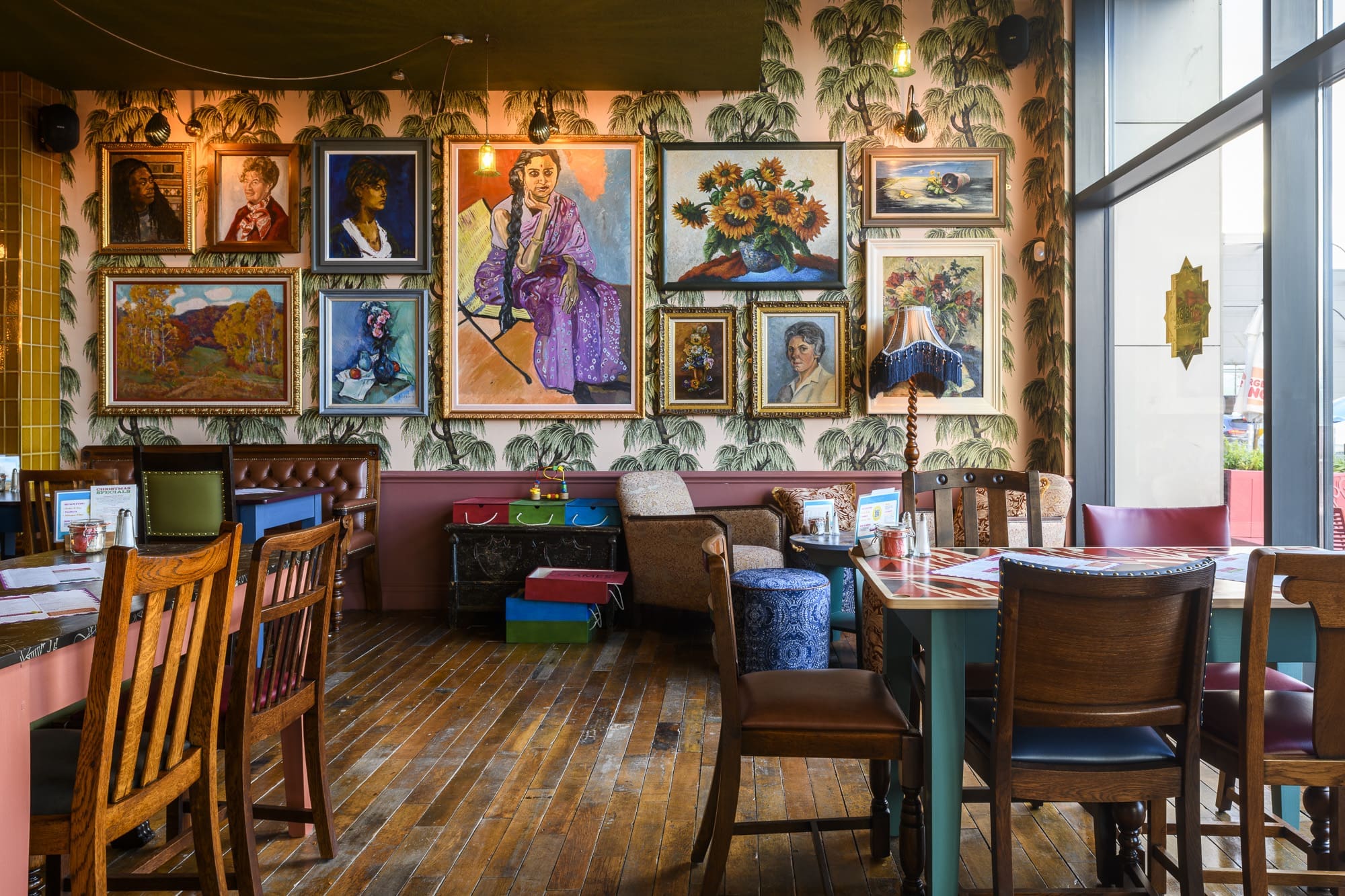 A cozy cafe with eclectic wooden furniture, vivid patterned wallpaper, and a gallery wall filled with colorful framed paintings—portraits, flowers, and even a coltello art piece. Natural light streams in through large windows on the right.