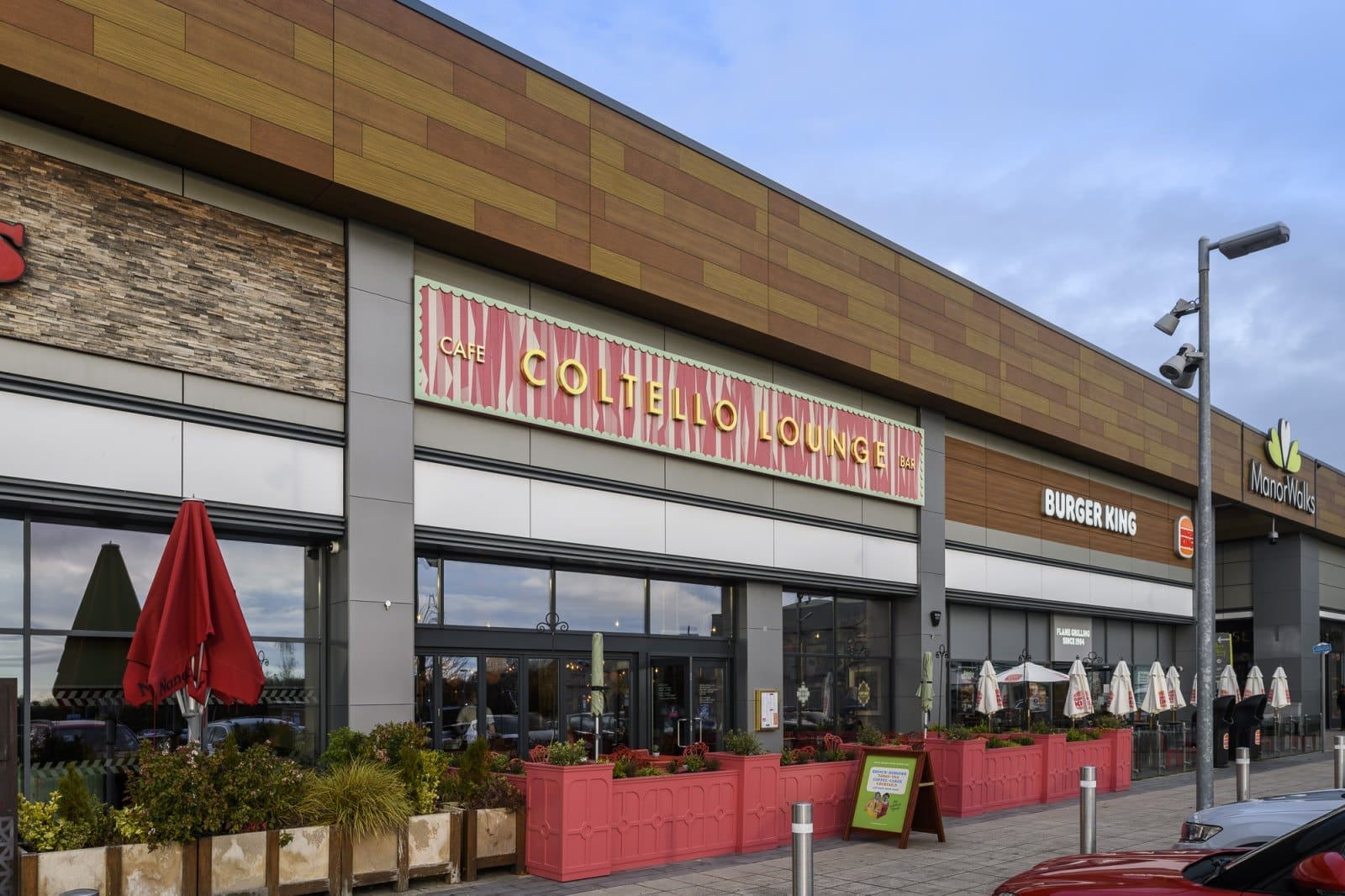 Outdoor view of a shopping center with Coltello Lounge café in the center, Burger King to the right, and outdoor seating areas featuring red planters and umbrellas along the walkway. Enjoy a relaxing atmosphere at Coltello under a cloudy sky.