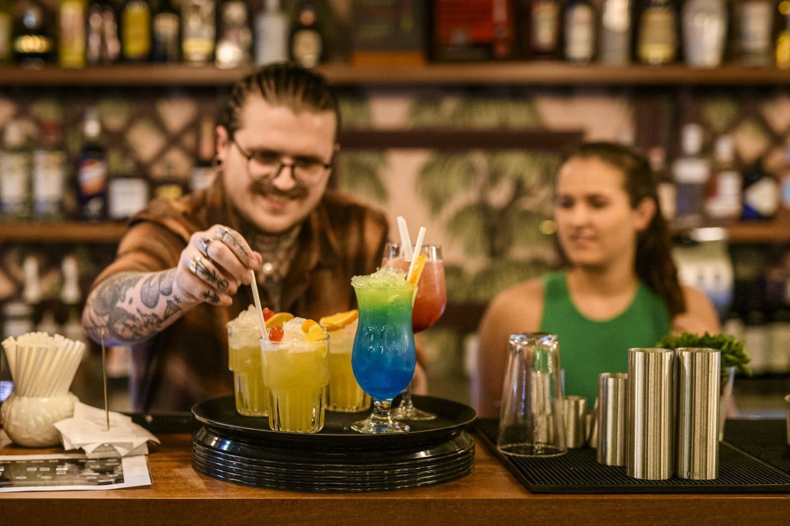 A bartender with tattoos arranges colorful cocktails on a tray at a bar, a coltello and other bar tools visible nearby, while a woman in a green top watches from behind the counter. Bottles line the background shelves.