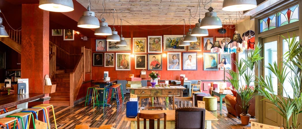 A cozy, colorful café in Como with eclectic chairs and tables, potted plants, and hanging lights. Portraits and art line the red walls. A staircase is visible in the background as sunlight streams in from large windows.
