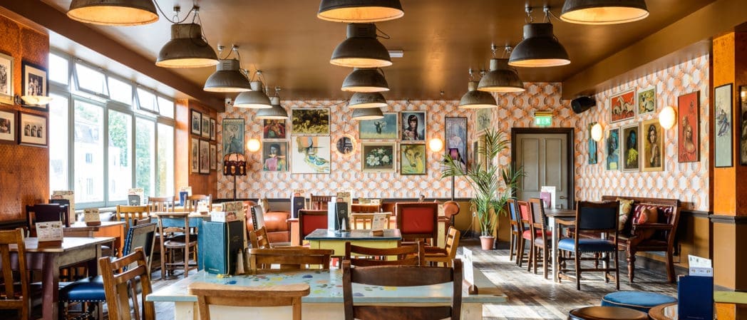 A cozy, brightly lit restaurant in Como with eclectic wooden tables and chairs, patterned wallpaper, hanging lights, framed artwork on the walls, and large windows letting in natural light.
