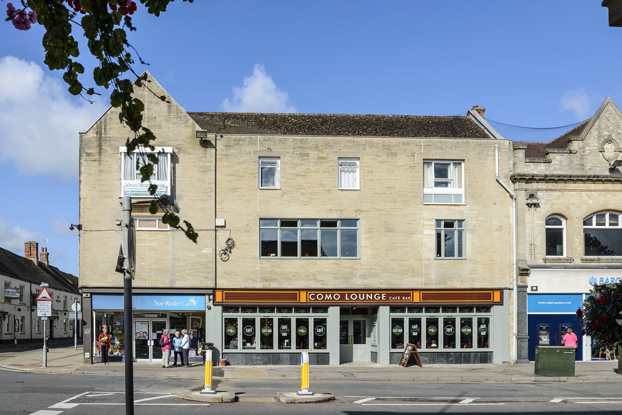 A street view of a beige stone building with large windows, home to the Como Lounge café bar on the ground floor. To the left, Sue Ryder’s blue shop stands out. Several people stroll along the sidewalk, adding life to this Como scene.