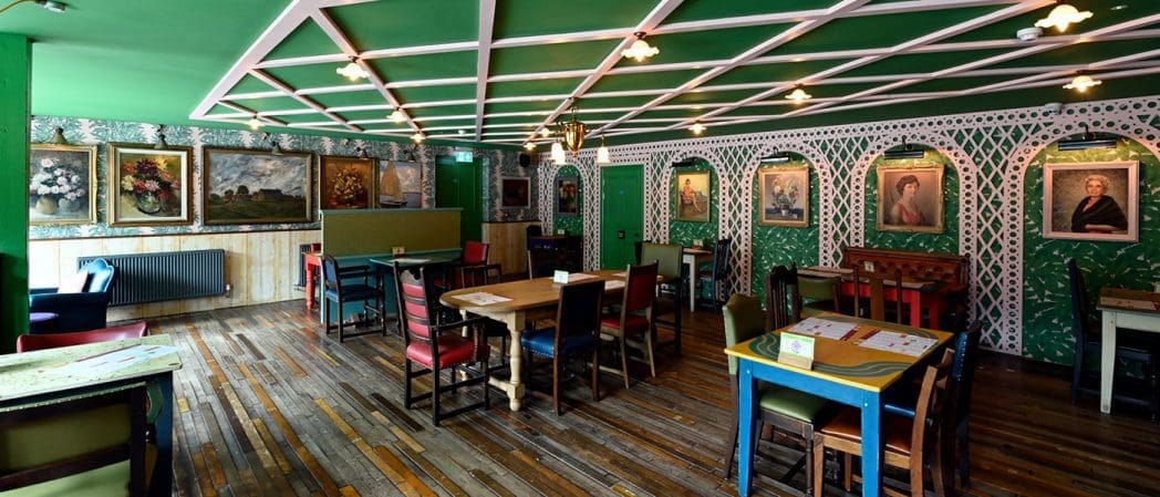 A cozy, eclectic restaurant dining room with mismatched chairs, green walls, wooden floors, and concho accents among the various framed paintings, creating a vintage atmosphere.