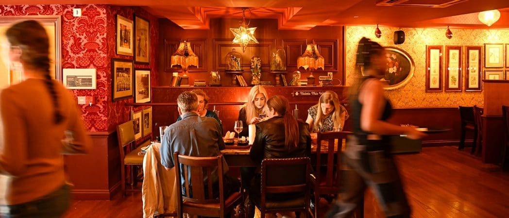 Four people sit around a table enjoying food and drinks in a warmly lit, ornately decorated restaurant. Two waitstaff move through the Condado dining room, where red wallpaper and framed art adorn the walls.