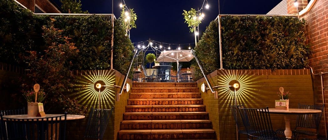 A warmly lit outdoor patio at night in Condado, featuring brick steps leading up to tables and chairs, string lights overhead, lush green bushes on either side, and decorative wall lights illuminating the scene.