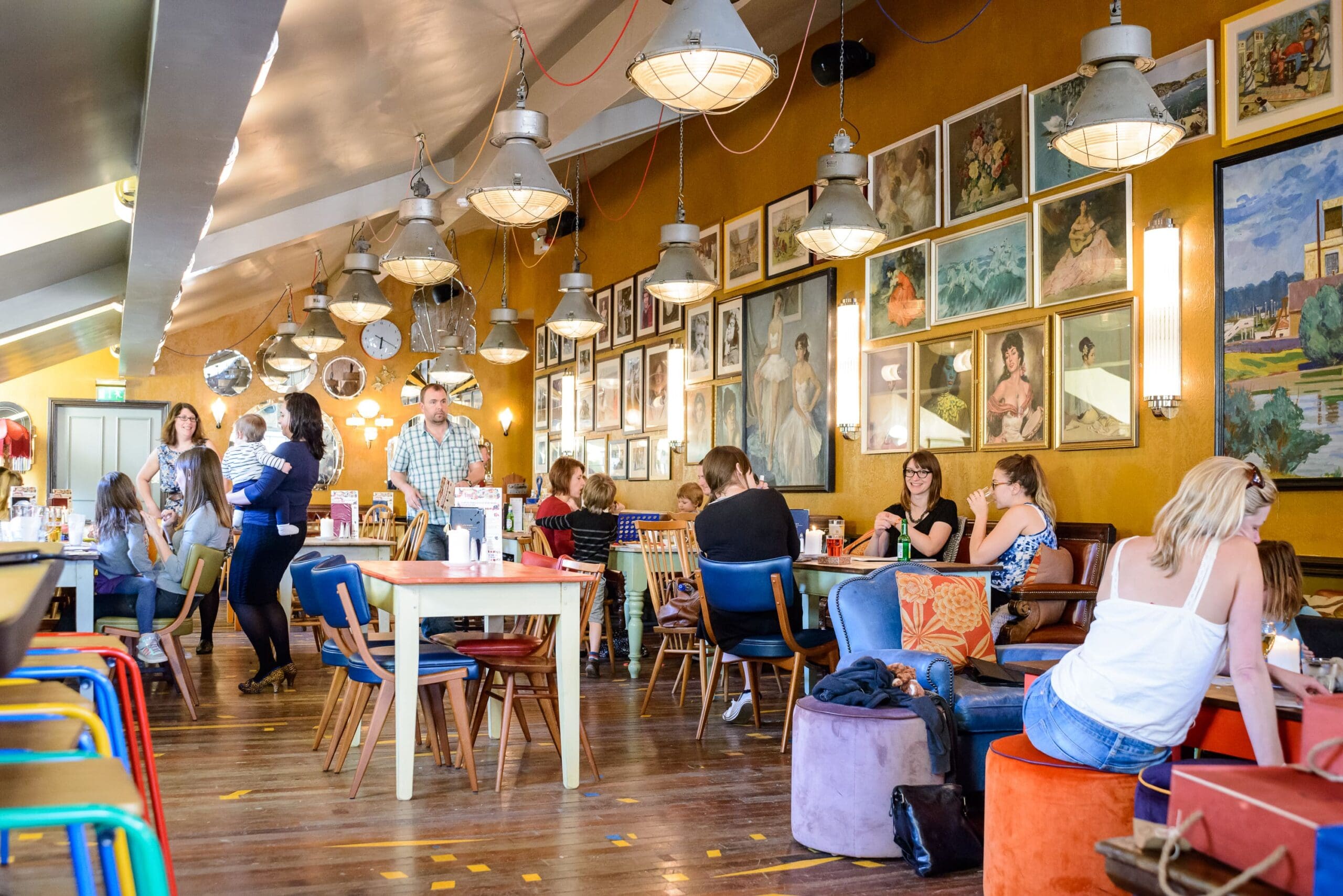 A busy, eclectic café with people dining and chatting at colorful tables and chairs. Framed artwork covers the yellow walls, while industrial-style lamps hang from the ceiling. Enjoy lively conversation over a delicious plate of Cordero in this casual atmosphere.