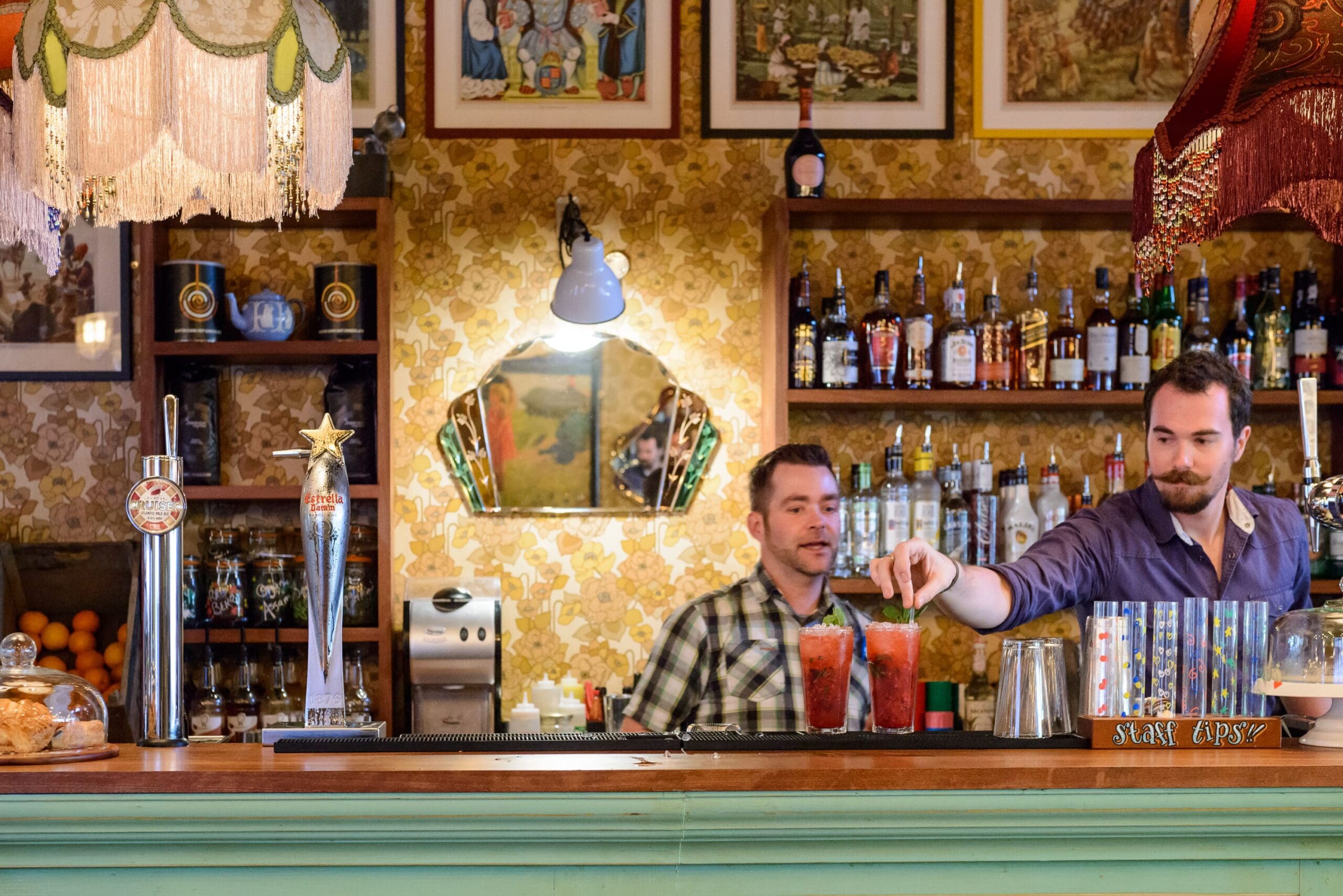 Two bartenders prepare and serve colorful Cordero cocktails behind a vintage-style bar, decorated with ornate lamps, patterned wallpaper, framed artwork, and shelves filled with bottles.