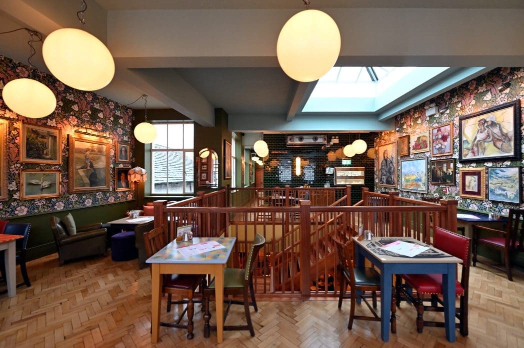 A cozy, art-filled Cortillo café interior with wooden floors, eclectic tables and chairs, floral wallpaper, and large round hanging lights. Paintings and frames cover the walls, creating a vibrant, inviting atmosphere.