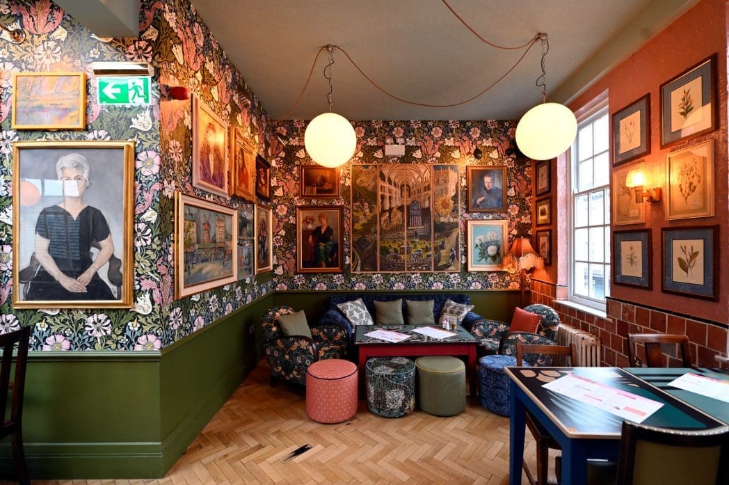 A cozy, eclectic Cortillo lounge with floral wallpaper, various framed artworks, round hanging lights, colorful chairs, couches, and ottomans, a red coffee table, and a wooden floor with a mix of vintage and modern decor.