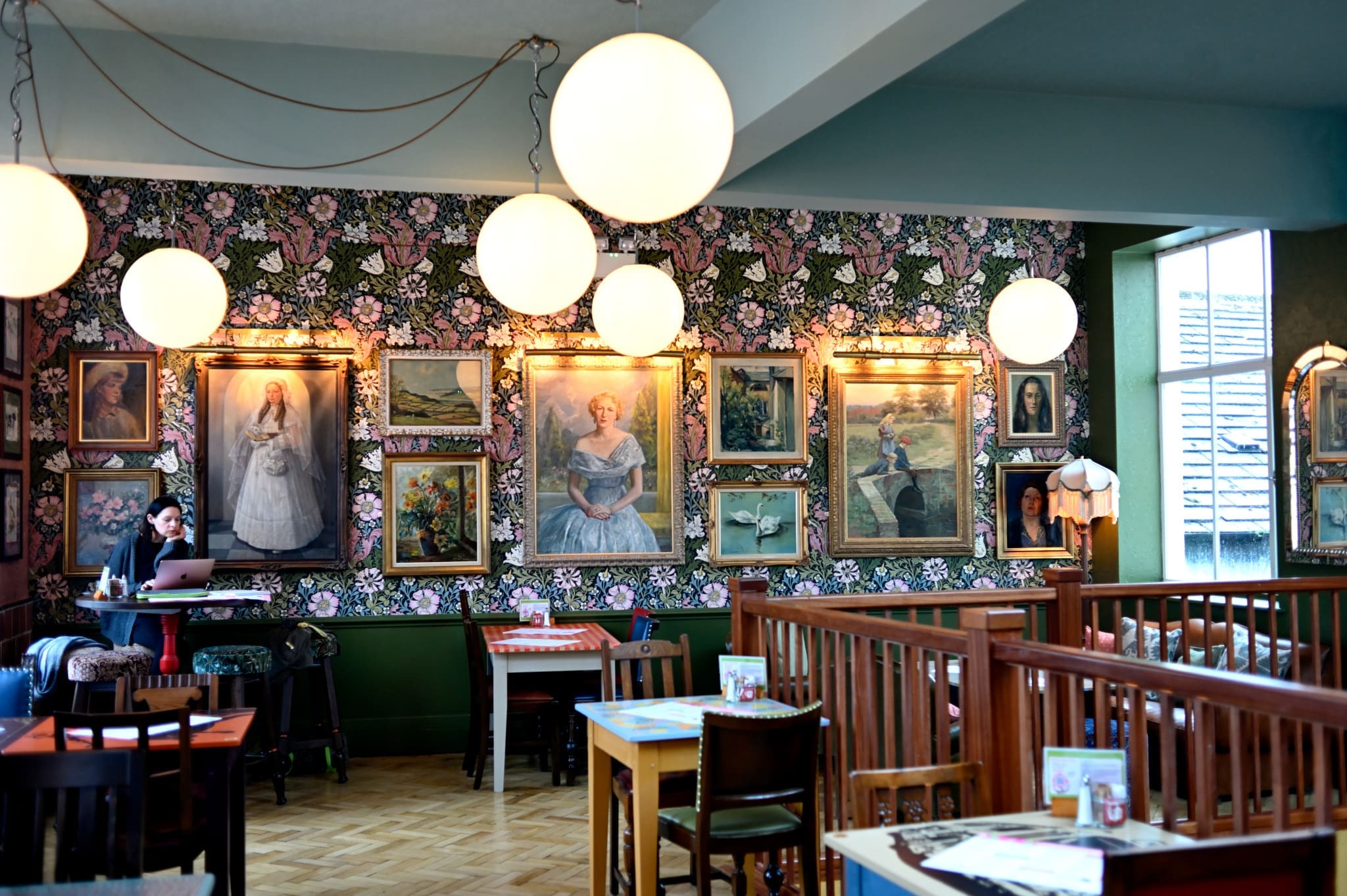 A cozy café with floral wallpaper, round hanging lights, and many framed portraits on the wall. Wooden tables and chairs are arranged throughout the space, creating a warm Cortillo vibe as a person works on a laptop in the corner.