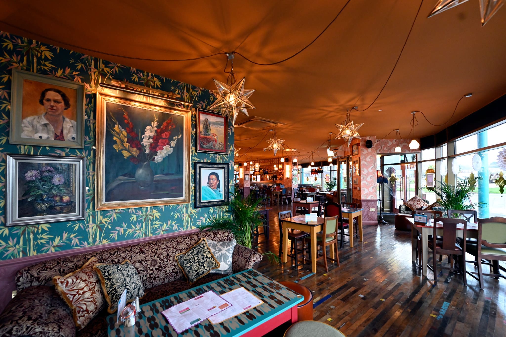 A cozy, vibrant Costero restaurant interior features patterned wallpaper, framed portraits, plush sofas, wooden tables, star-shaped pendant lights, and large windows that flood the space with natural light.