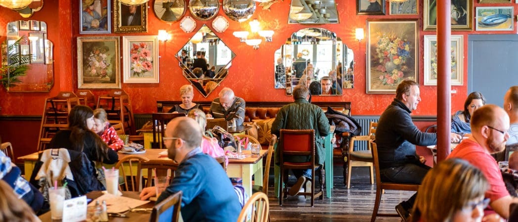 People sit at tables enjoying food and conversation in a lively, colorful Cultivo café decorated with framed art, mirrors, and red walls. The scene is busy and inviting, creating a warm atmosphere.
